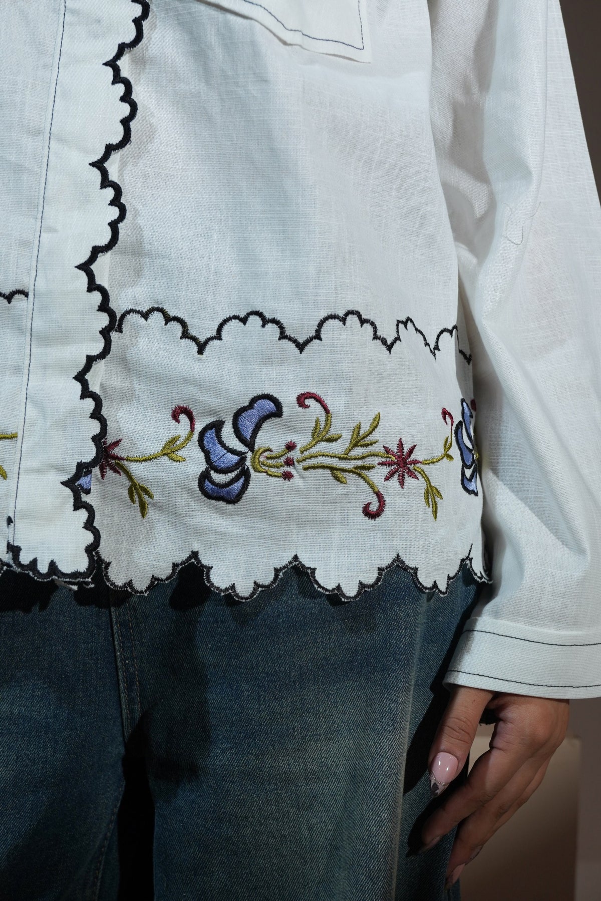 A man wearing a white long-sleeve button-up casual wear shirt with red embroidered text and a floral lace hem, paired with wide-leg blue jeans and red socks, sitting on a beige block against a solid beige background.