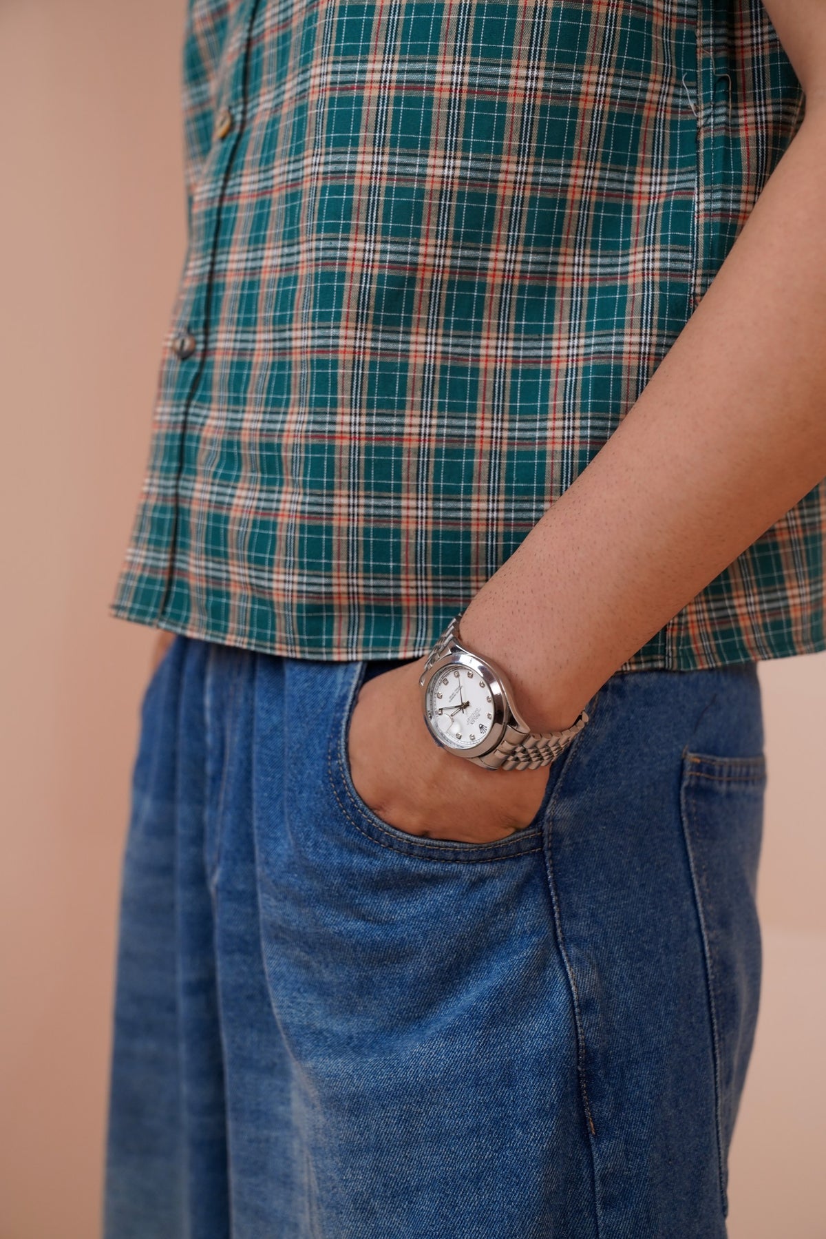 A close-up of a person wearing a green and orange plaid shirt, blue denim jeans, and a silver-toned metal link watch with a white face.