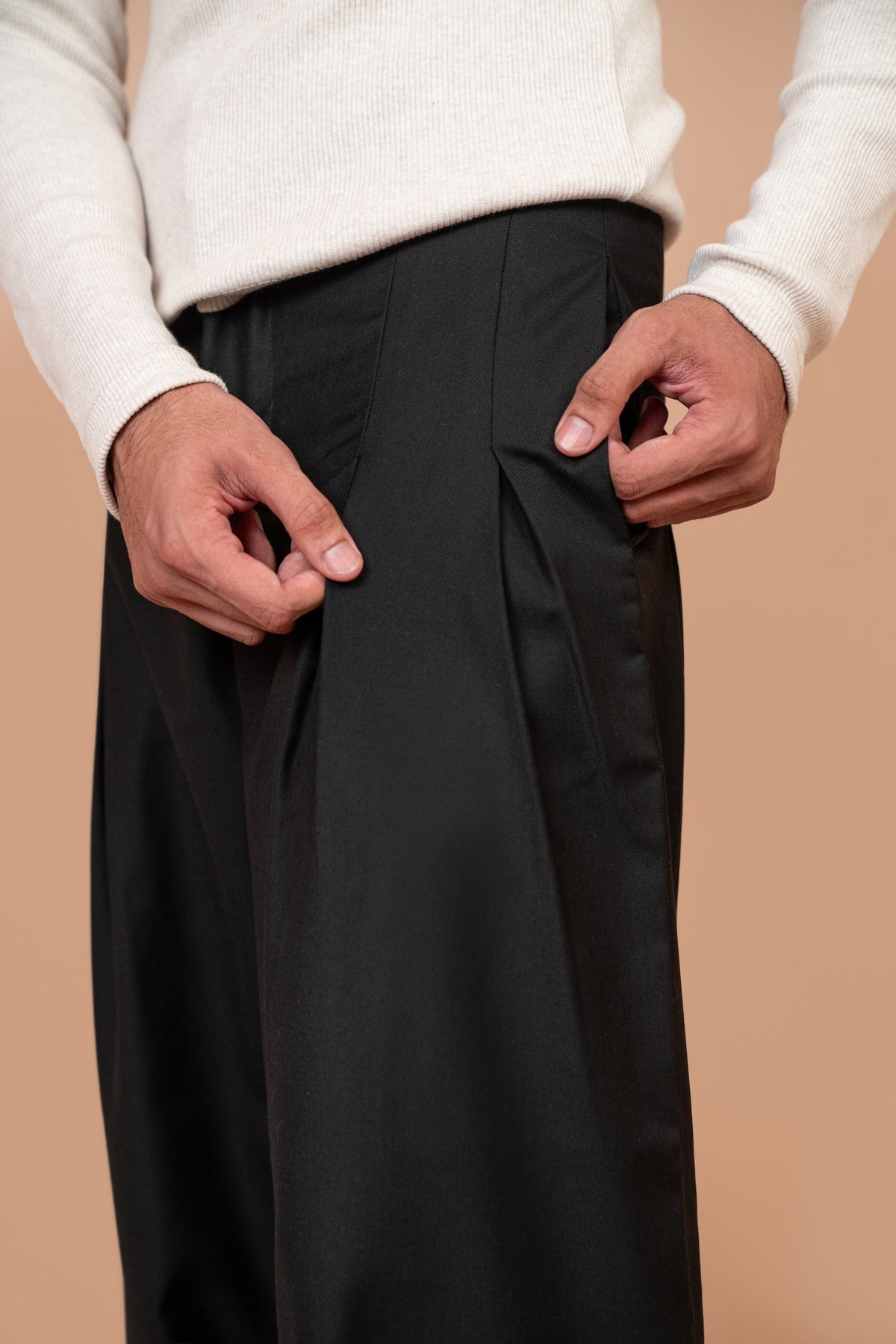 A close-up of a person wearing black wide-leg trousers and a cream ribbed sweater, with both hands adjusting the side pocket of the trousers, set against a solid warm beige background.
