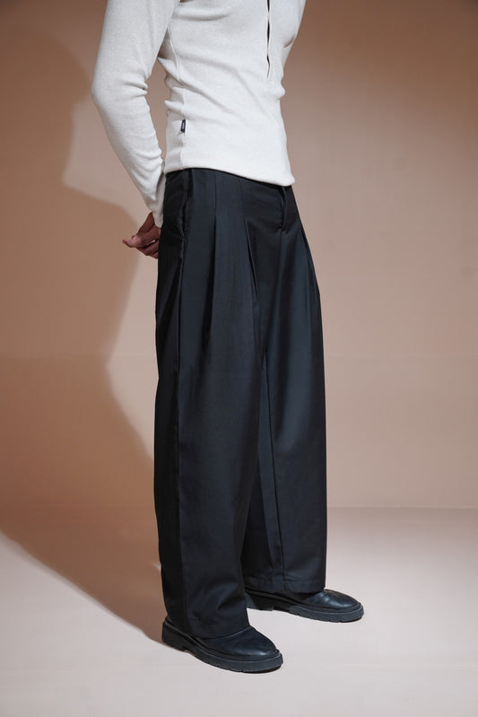 A model wearing wide-leg black trousers, a white long-sleeve top, and black leather shoes, standing against a warm beige studio backdrop.