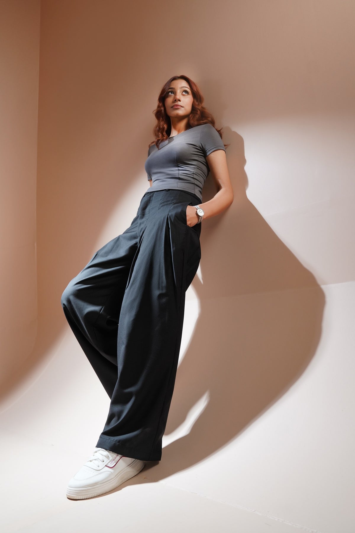 A woman posing in a studio setting wearing a gray short-sleeve crop top, dark charcoal wide-leg trousers, and white sneakers, leaning against a beige wall with dramatic shadows.