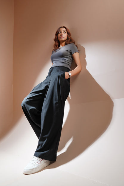 A woman posing in a studio setting wearing a gray short-sleeve crop top, dark charcoal wide-leg trousers, and white sneakers, leaning against a beige wall with dramatic shadows.