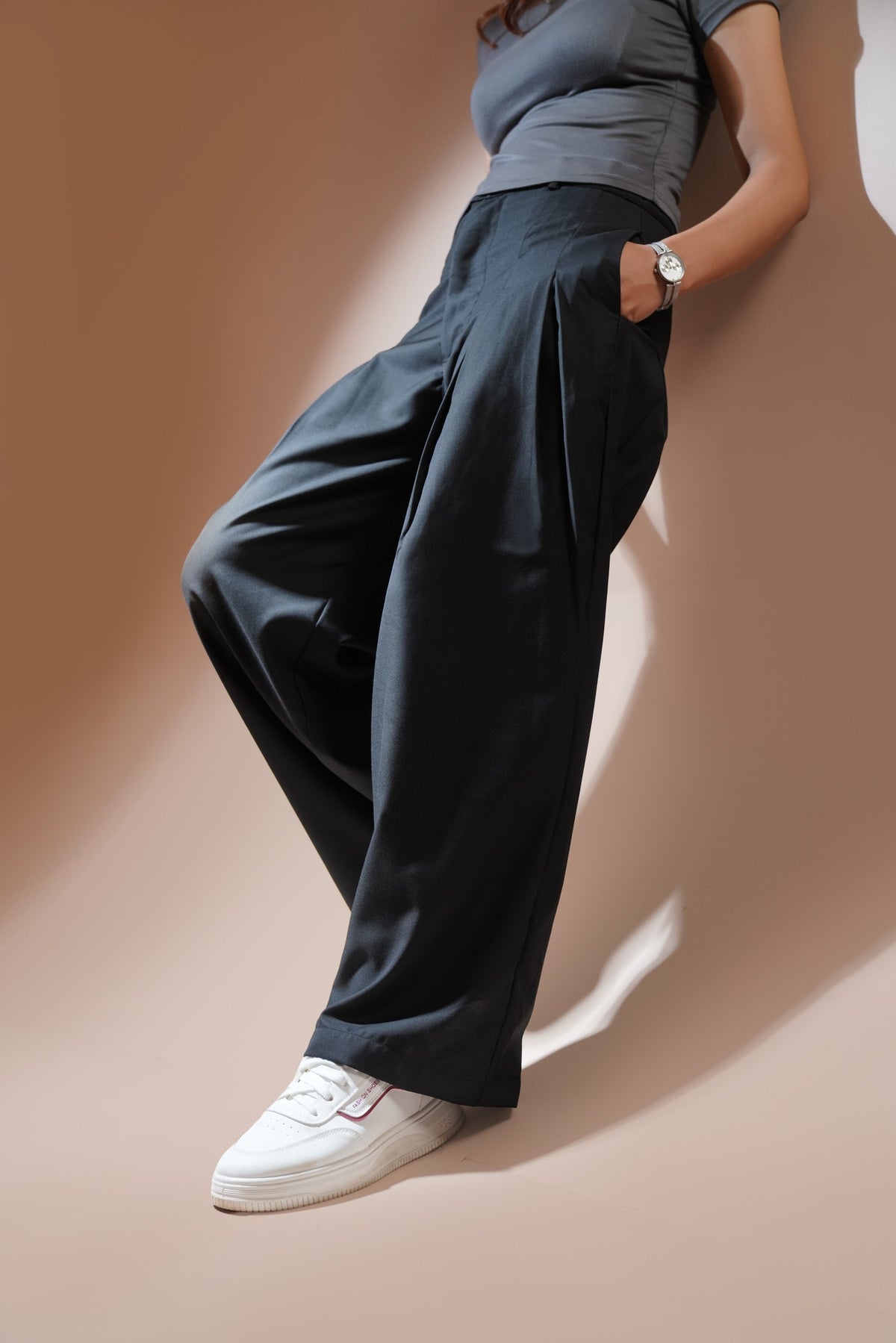 A model wearing black wide-leg pleated trousers, a gray t-shirt, and white sneakers, leaning against a warm beige wall with dramatic shadows.