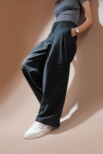 A model wearing black wide-leg pleated trousers, a gray t-shirt, and white sneakers, leaning against a warm beige wall with dramatic shadows.