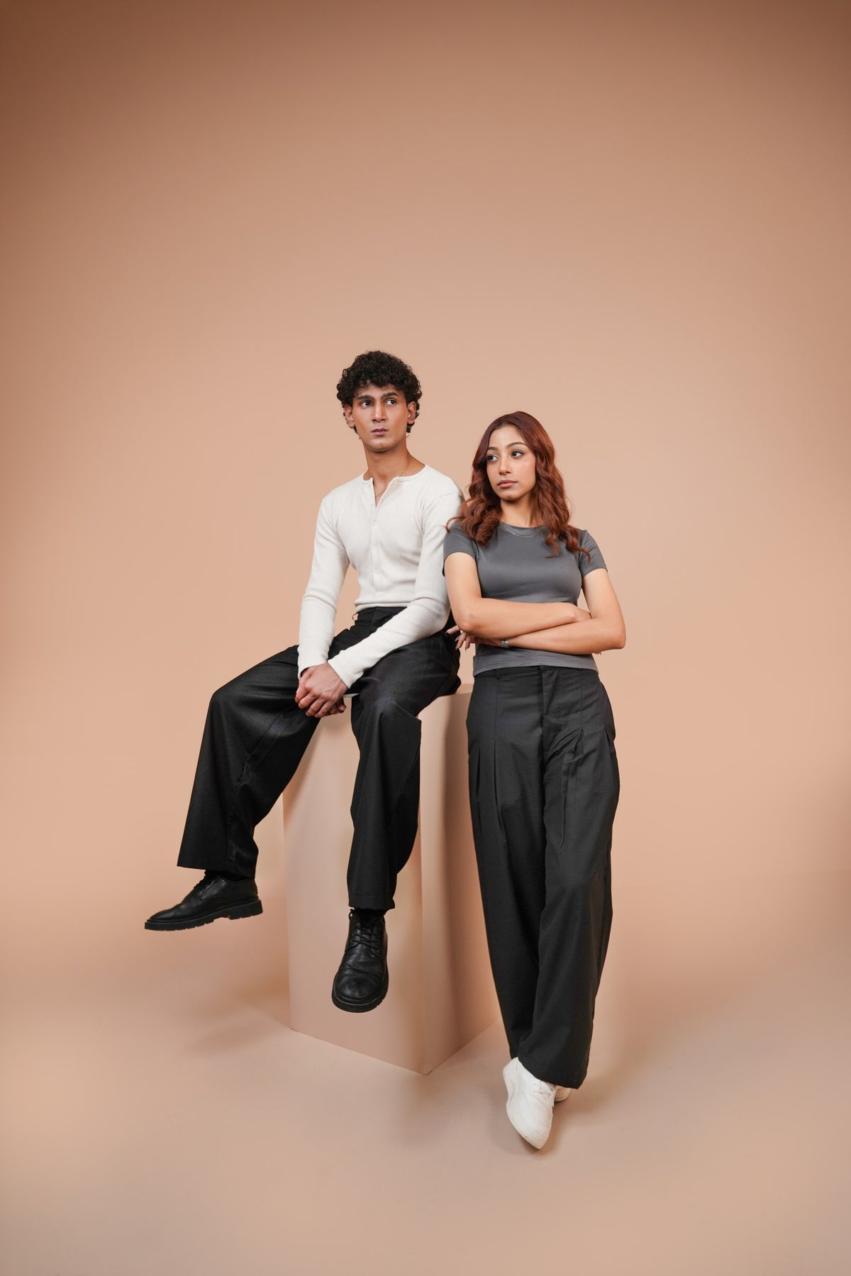 Two models posing in minimalist clothing against a solid peach studio backdrop. The male model sits on a matching pedestal wearing a white long-sleeve henley shirt and black wide-leg trousers, while the female model stands beside him in a gray short-sleeve top and black wide-leg trousers.