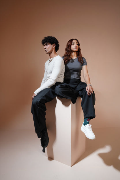A man and a woman pose back-to-back sitting on a tall beige pedestal against a warm beige studio backdrop. The man wears a white long-sleeve top and black pants, while the woman wears a dark gray fitted top, black pants, and white sneakers.