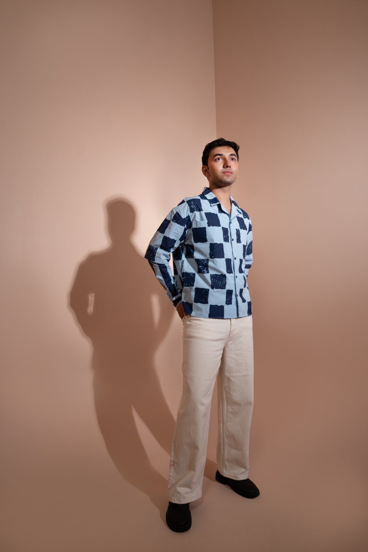 A man stands in a studio corner wearing a blue and white checkered unisex shirt, off-white trousers, and black shoes. A strong shadow is cast on the beige wall behind him.