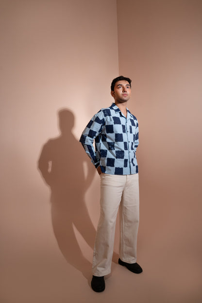 A man stands in a studio corner wearing a blue and white checkered unisex shirt, off-white trousers, and black shoes. A strong shadow is cast on the beige wall behind him.
