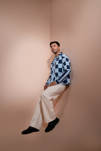 A man wearing a blue and white checkered unisex shirt, cream wide-leg trousers, and black shoes, leaning against a warm beige corner wall.