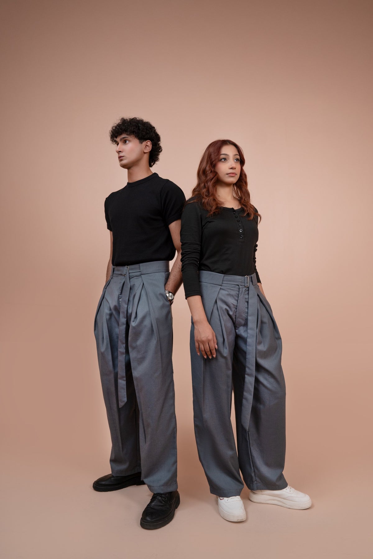 A man and a woman standing back-to-back wearing matching high-waisted, wide-leg gray trousers, black long-sleeve tops, and casual shoes against a solid peach-colored studio background.
