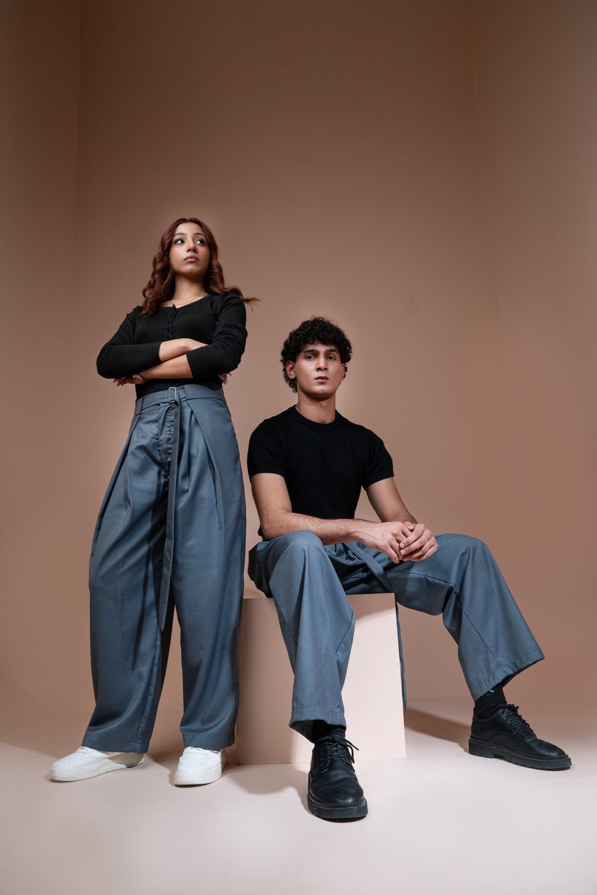 A man and a woman pose in matching black crewneck t-shirts and wide-leg trousers, set against a minimalist warm beige studio backdrop.