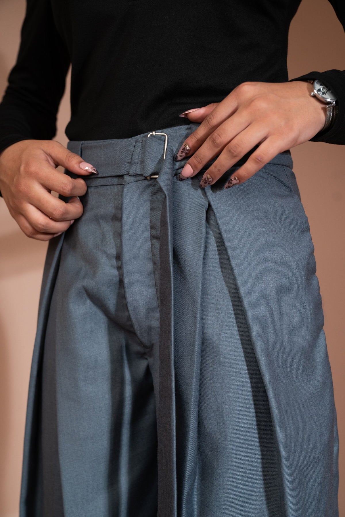 Close-up of a person wearing high-waisted slate blue pleated trousers with a matching belt and silver buckle, paired with a black long-sleeve top.