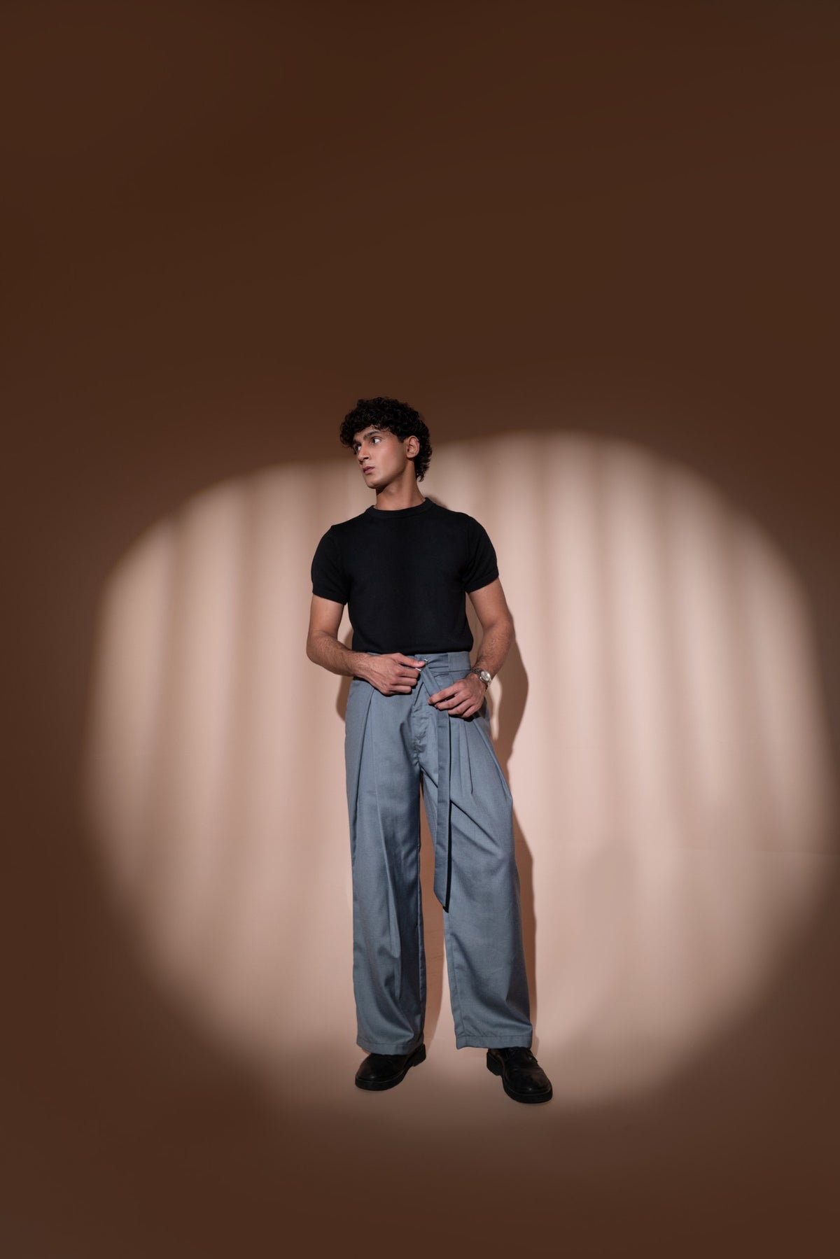 A man stands in a studio wearing a fitted black short-sleeve t-shirt and light gray wide-leg trousers, posed against a warm brown backdrop with a spotlight effect behind him.