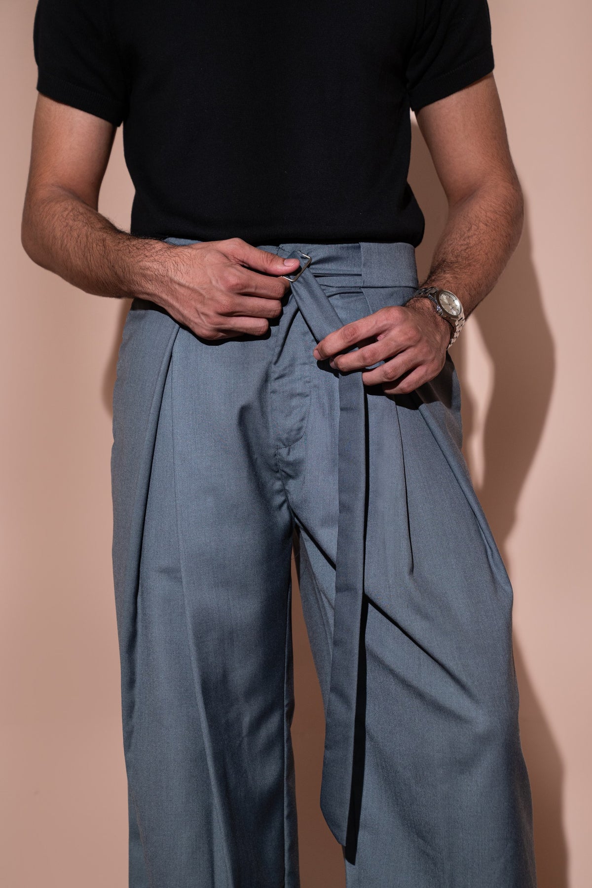 A close-up of a person wearing slate blue wide-leg trousers with a self-tie waist belt, paired with a black short-sleeve top against a solid beige background.
