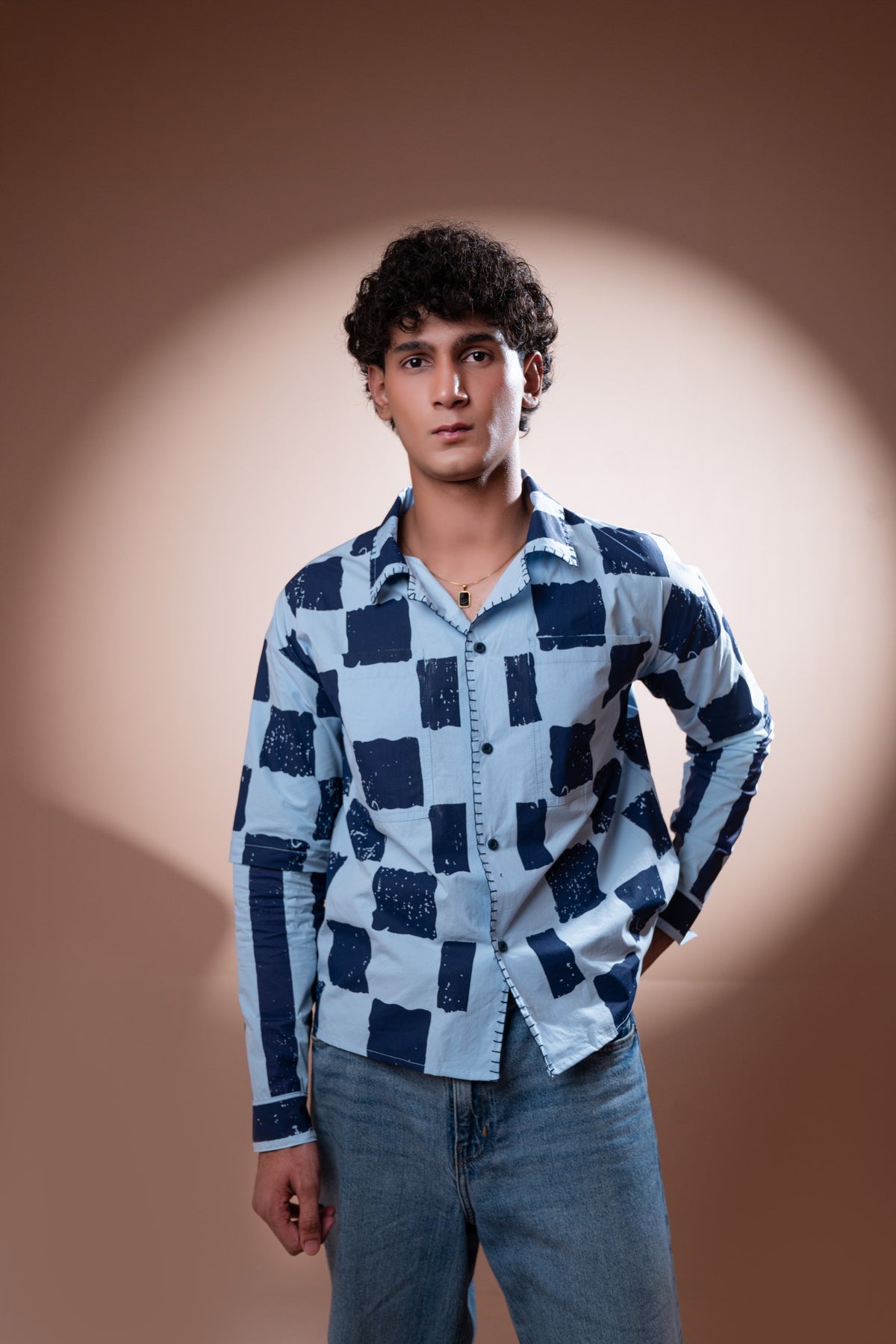 A young man with curly dark hair stands against a warm brown studio backdrop with a spotlight effect, wearing a light blue and navy checkered unisex shirt and blue jeans.