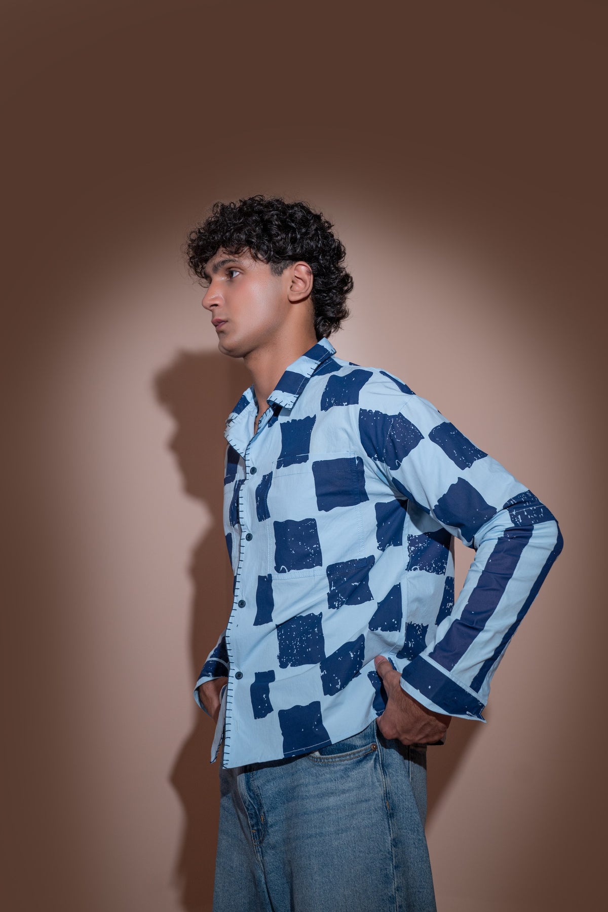 A young man with curly black hair stands in profile against a warm brown studio background, wearing a light blue and navy checkered long-sleeve unisex shirt and blue jeans.