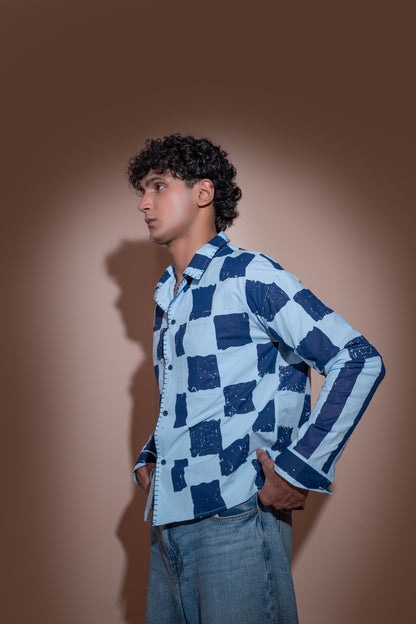 A young man with curly black hair stands in profile against a warm brown studio background, wearing a light blue and navy checkered long-sleeve unisex shirt and blue jeans.