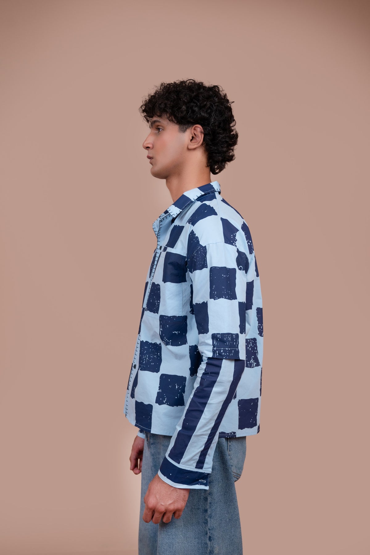 A man in profile wearing a light blue and navy checkered shirt with textured squares and striped details on the sleeves, paired with light blue jeans, set against a solid warm beige background.