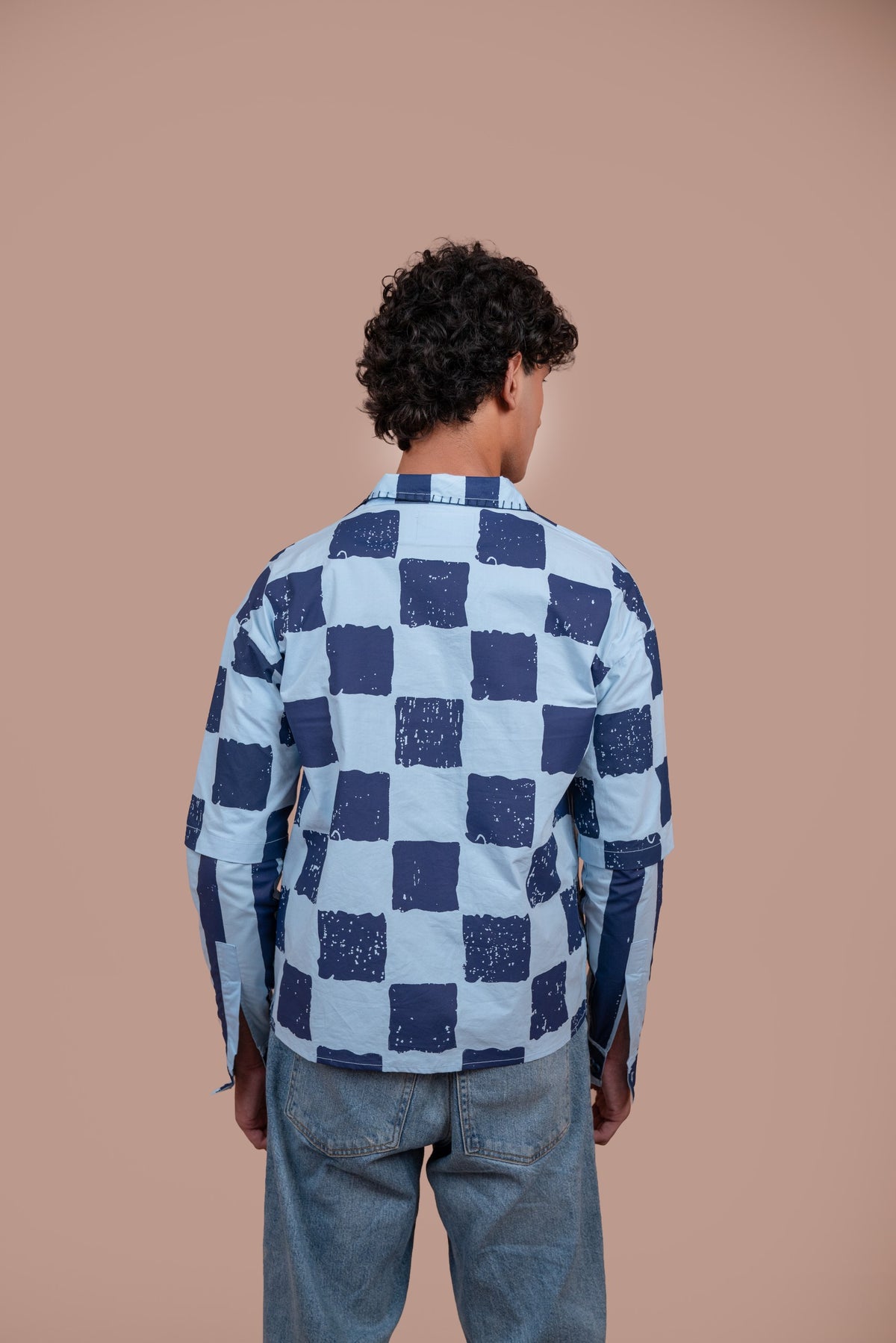 A man viewed from behind wearing a light blue and navy blue checkered shirt and light blue jeans, standing against a solid warm beige background.