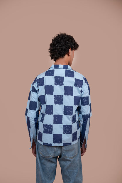 A man viewed from behind wearing a light blue and navy blue checkered shirt and light blue jeans, standing against a solid warm beige background.