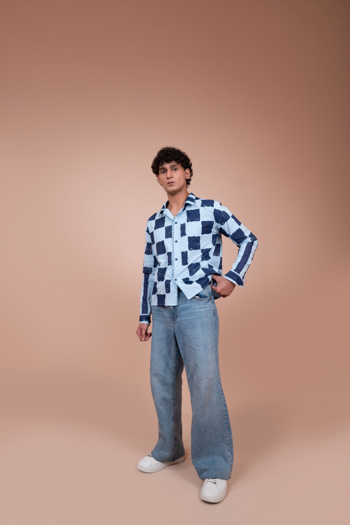 A man modeling a long-sleeve unisex shirt with a navy and white checkerboard pattern, paired with light-wash wide-leg jeans and white sneakers, set against a solid warm beige studio background.