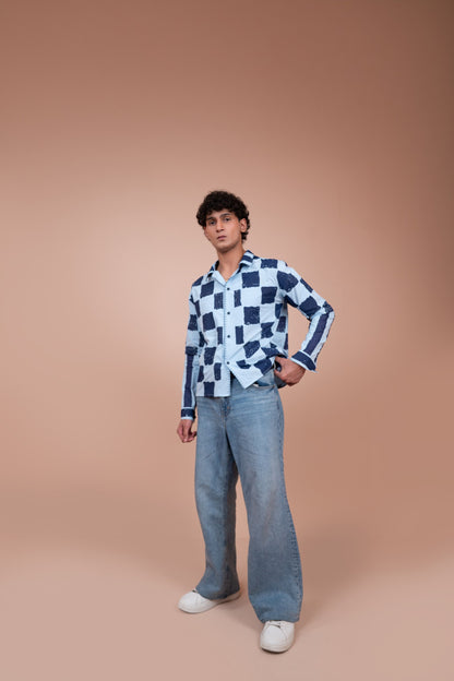 A man modeling a long-sleeve unisex shirt with a navy and white checkerboard pattern, paired with light-wash wide-leg jeans and white sneakers, set against a solid warm beige studio background.