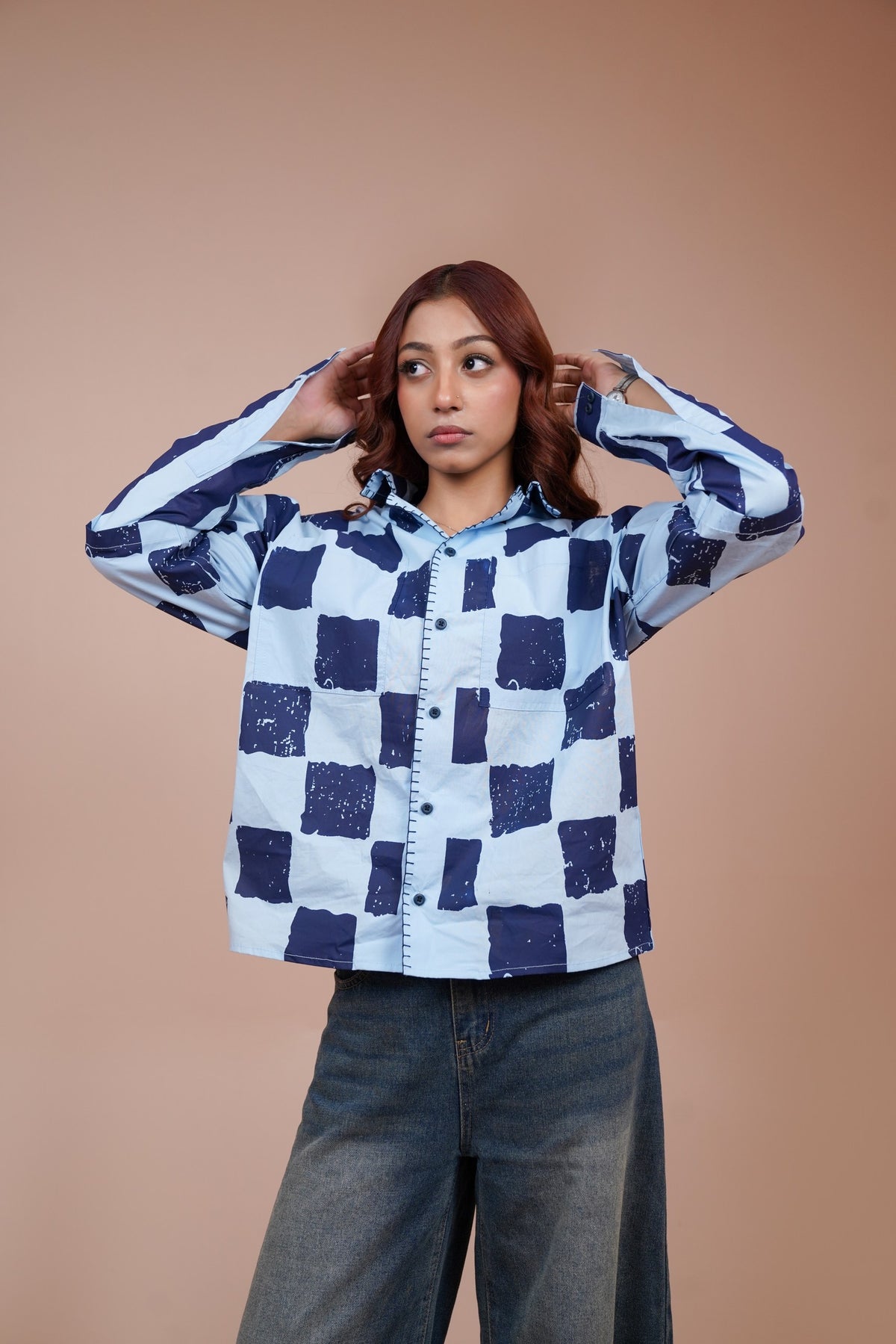 A woman modeling a light blue and navy checkered unisex shirt with wide-leg blue jeans, posing against a solid warm beige studio background.