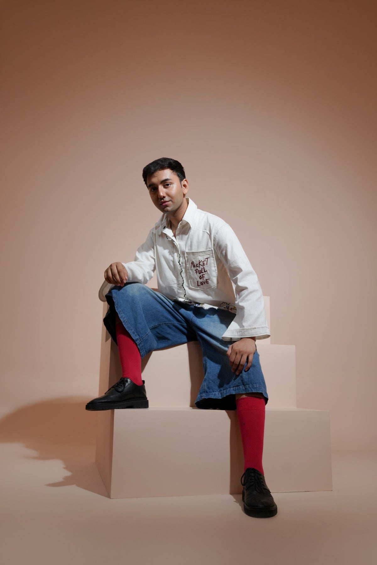 A man sits on a geometric platform wearing a white embroidered casualwear shirt blue denim shorts, bright red socks, and black shoes against a warm beige studio backdrop.