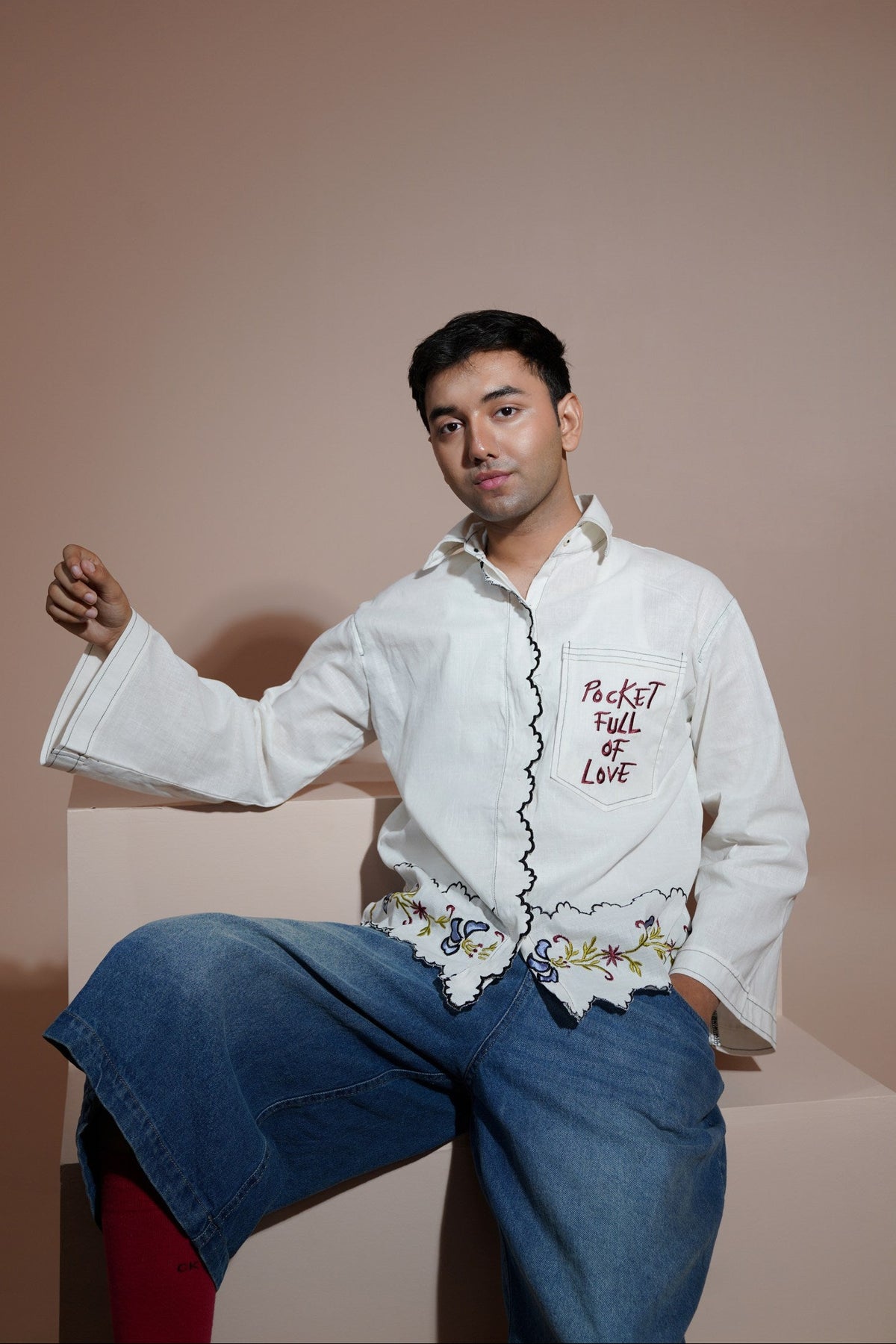 A man wearing a white long-sleeve button-up casual wear shirt with red embroidered text and a floral lace hem, paired with wide-leg blue jeans and red socks, sitting on a beige block against a solid beige background.