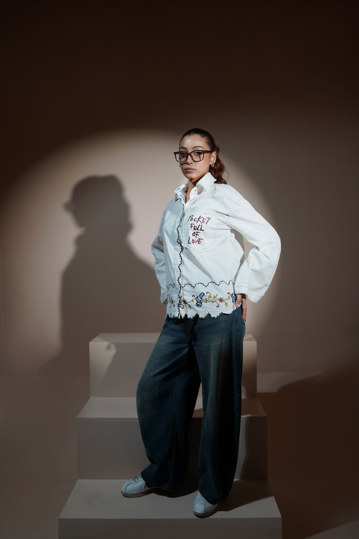 A man wearing a white long-sleeve button-up casual wear shirt with red embroidered text and a floral lace hem, paired with wide-leg blue jeans and red socks, sitting on a beige block against a solid beige background.