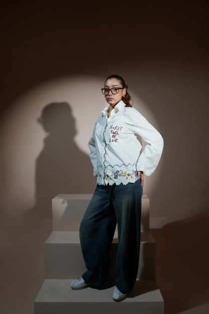 A man wearing a white long-sleeve button-up casual wear shirt with red embroidered text and a floral lace hem, paired with wide-leg blue jeans and red socks, sitting on a beige block against a solid beige background.