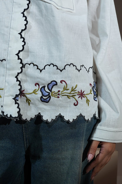 A man wearing a white long-sleeve button-up casual wear shirt with red embroidered text and a floral lace hem, paired with wide-leg blue jeans and red socks, sitting on a beige block against a solid beige background.