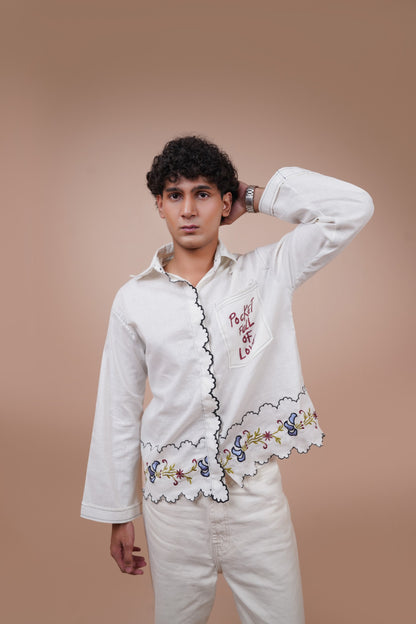A man wearing a white long-sleeve button-up casual wear shirt with red embroidered text and a floral lace hem, paired with wide-leg blue jeans and red socks, sitting on a beige block against a solid beige background.
