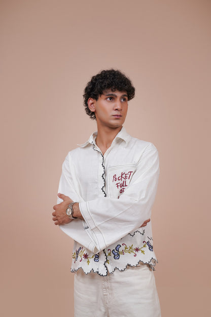A man wearing a white long-sleeve button-up casual wear shirt with red embroidered text and a floral lace hem, paired with wide-leg blue jeans and red socks, sitting on a beige block against a solid beige background.