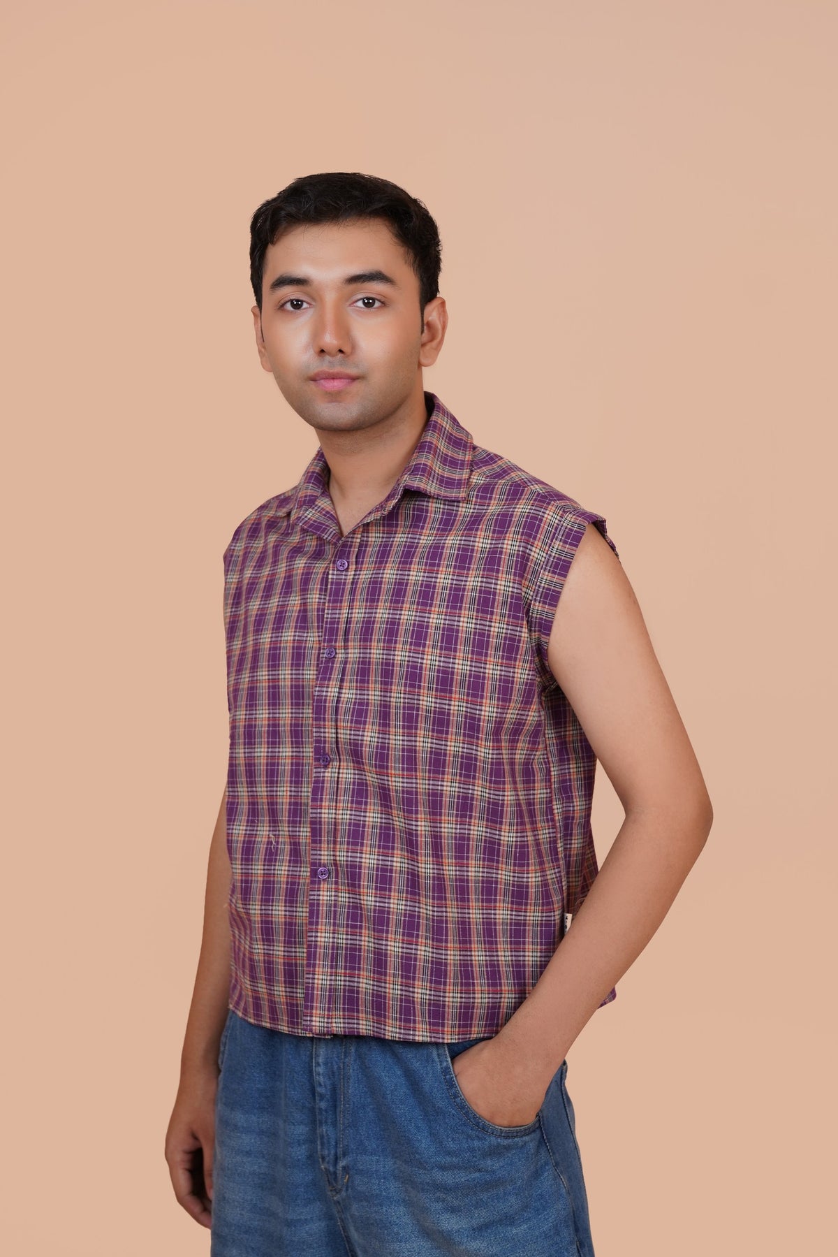 A man wearing a sleeveless purple and red plaid button-up shirt with blue jeans, standing against a solid beige background.