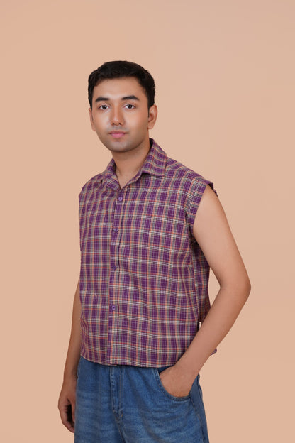 A man wearing a sleeveless purple and red plaid button-up shirt with blue jeans, standing against a solid beige background.