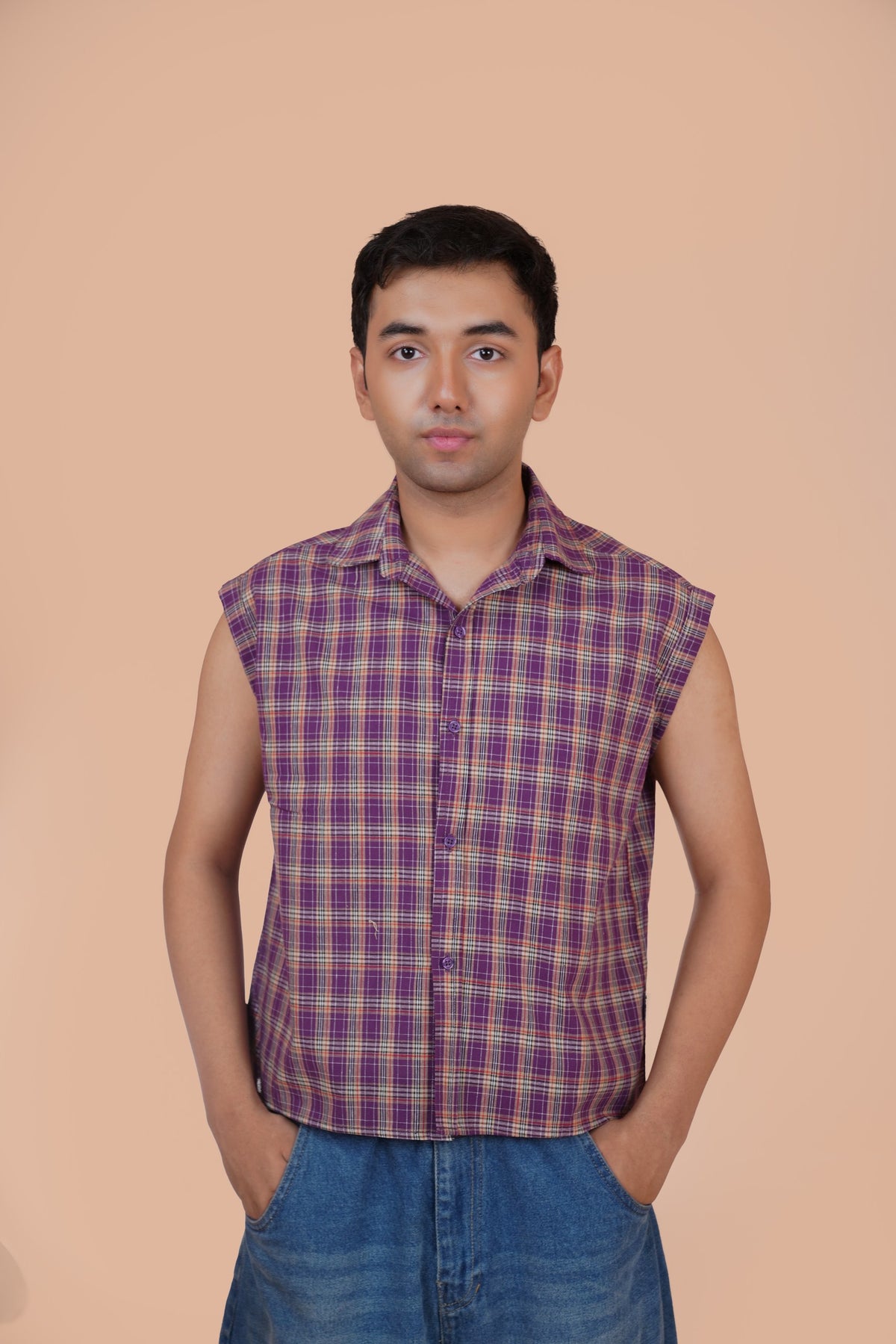 A man wearing a sleeveless purple and red plaid button-up shirt with blue jeans, standing against a solid peach-colored background.