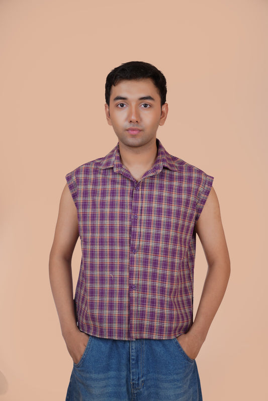 A man wearing a sleeveless purple and red plaid button-up shirt with blue jeans, standing against a solid peach-colored background.