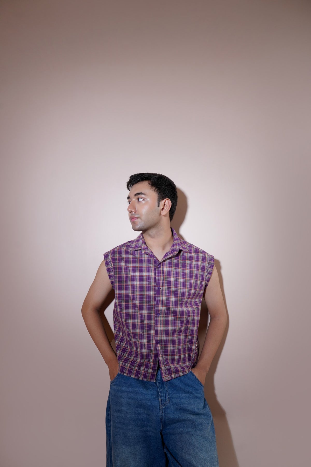 A man wearing a purple and blue plaid sleeveless button-up shirt and blue jeans, standing against a solid beige studio background.