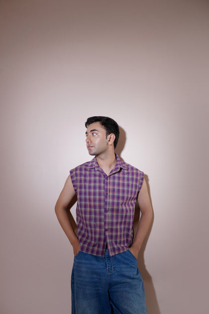 A man wearing a purple and blue plaid sleeveless button-up shirt and blue jeans, standing against a solid beige studio background.