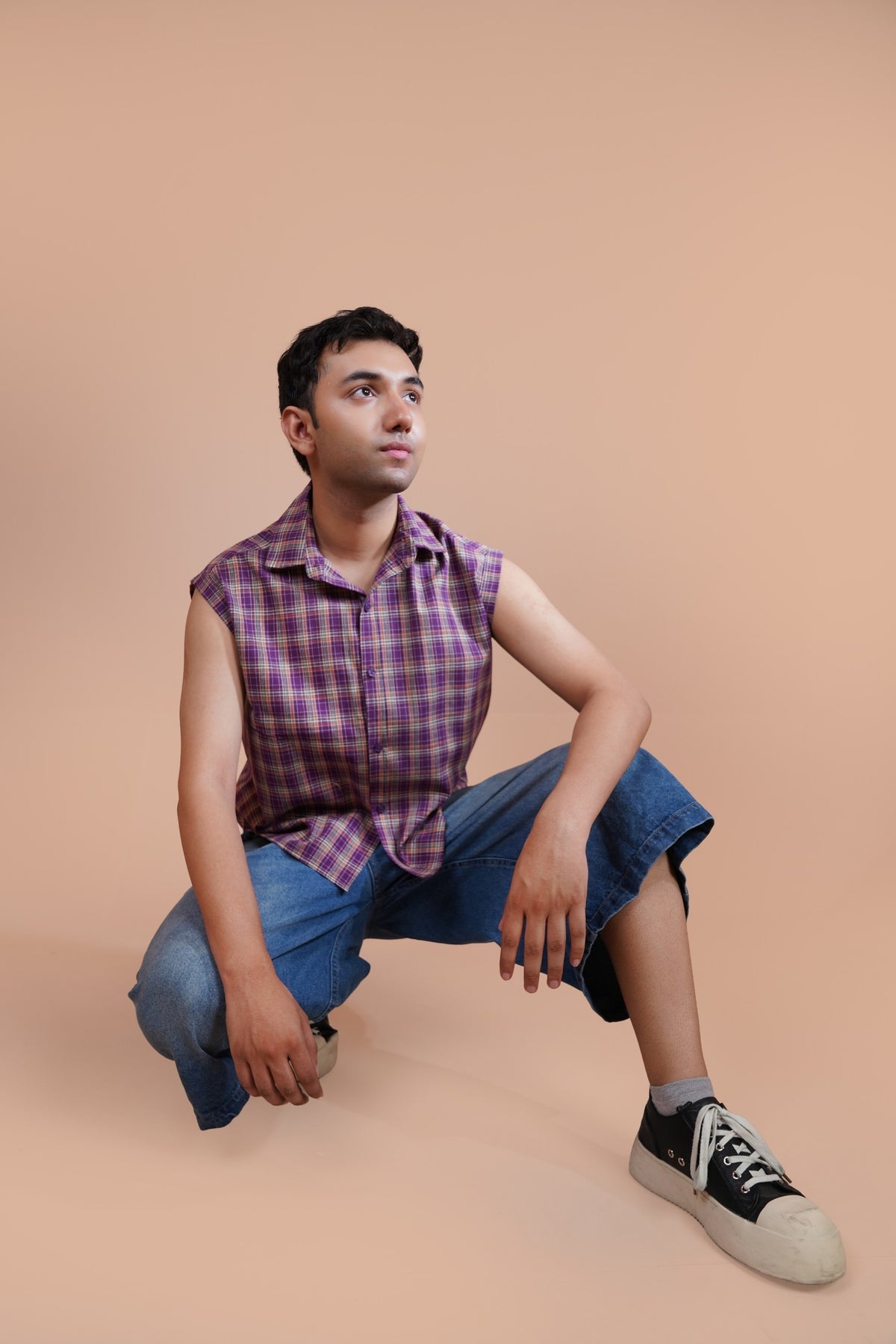 A man wearing a purple and pink plaid sleeveless button-up shirt, blue denim shorts, and black sneakers with white toe caps, crouching against a solid beige studio background.