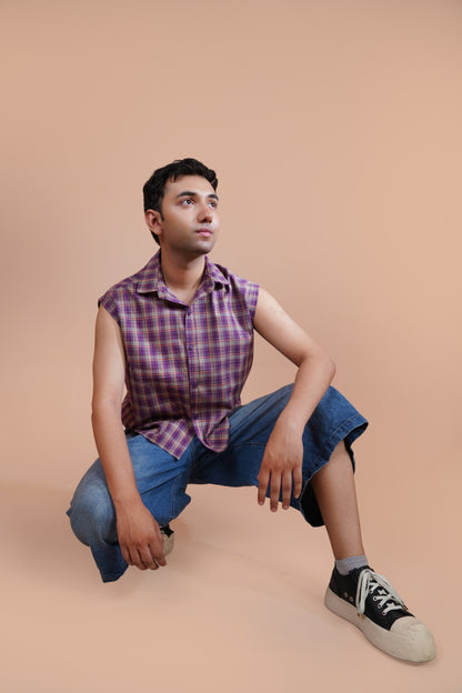 A man wearing a purple and pink plaid sleeveless button-up shirt, blue denim shorts, and black sneakers with white toe caps, crouching against a solid beige studio background.