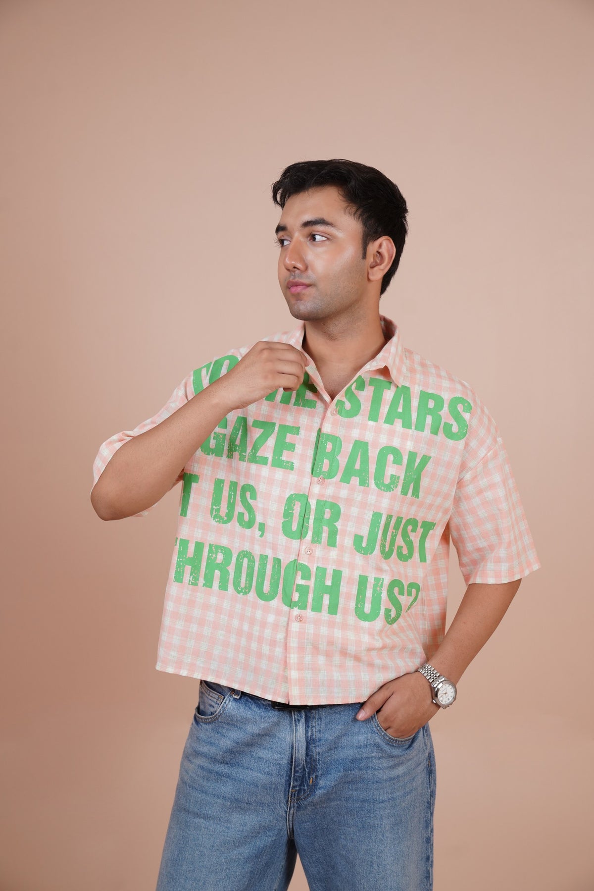 A man wearing a pink and white gingham short-sleeve-shirt with bold green text that reads 'LOOK AT THE STARS GAZE BACK AT US, OR JUST THROUGH US?' paired with blue jeans, posed against a solid beige studio background.