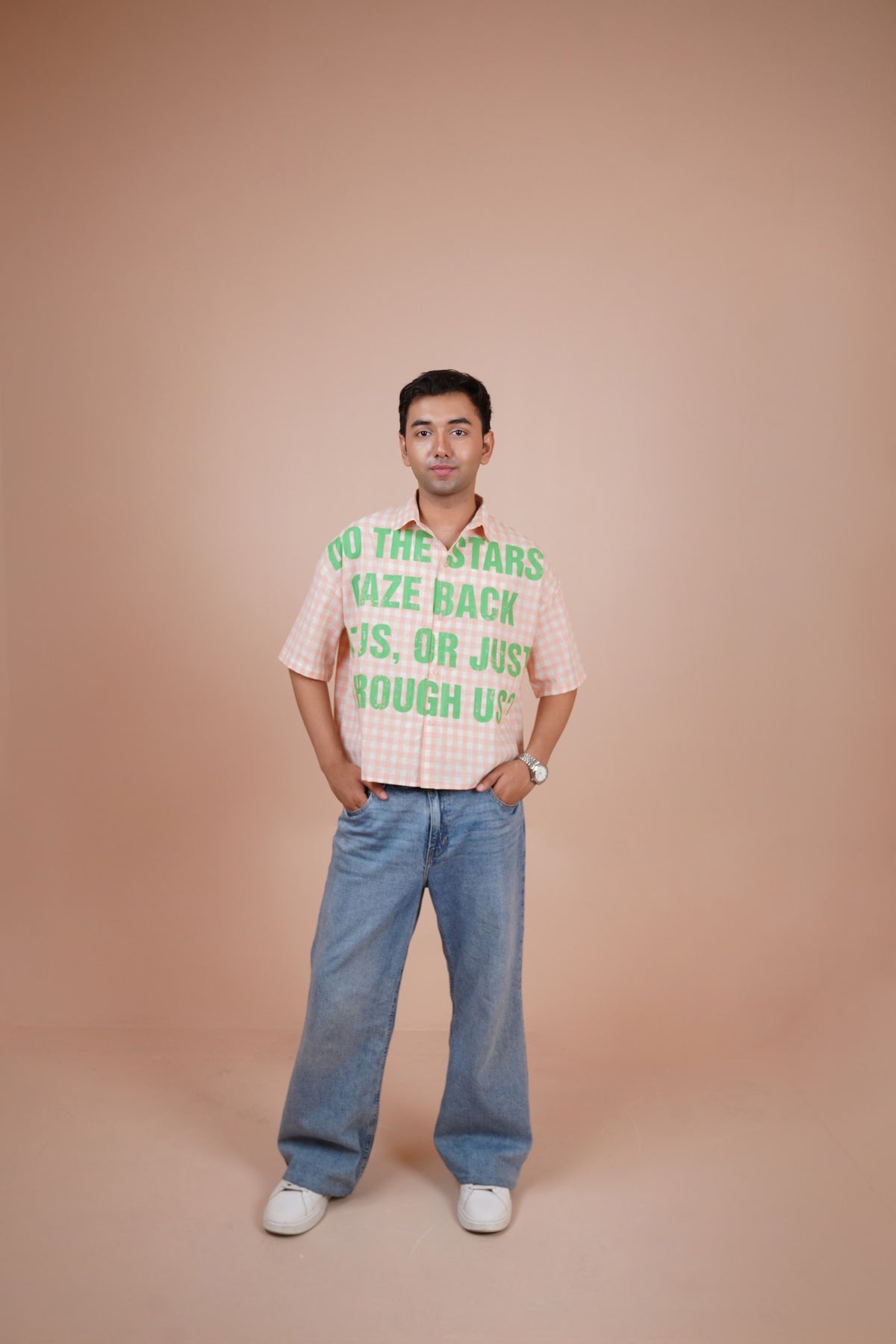 A man stands against a solid peach studio backdrop wearing a short-sleeve-shirt with green text that reads 'DO THE STARS WAZE BACK US, OR JUST ROUGH US', paired with wide-leg blue jeans and white sneakers.