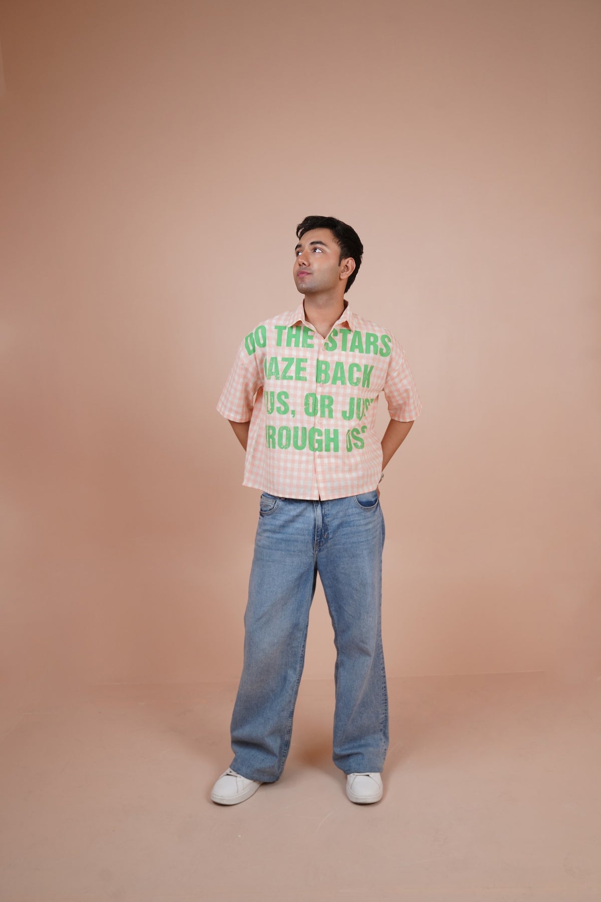 A man wearing a pink and white gingham short-sleeve-shirt with green text that reads 'DO THE STARS MAZE BACK US, OR JUST BOUGH S' paired with wide-leg blue jeans and white sneakers, standing against a solid peach-colored studio backdrop.