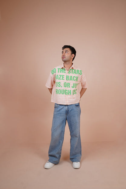 A man wearing a pink and white gingham short-sleeve-shirt with green text that reads 'DO THE STARS MAZE BACK US, OR JUST BOUGH S' paired with wide-leg blue jeans and white sneakers, standing against a solid peach-colored studio backdrop.