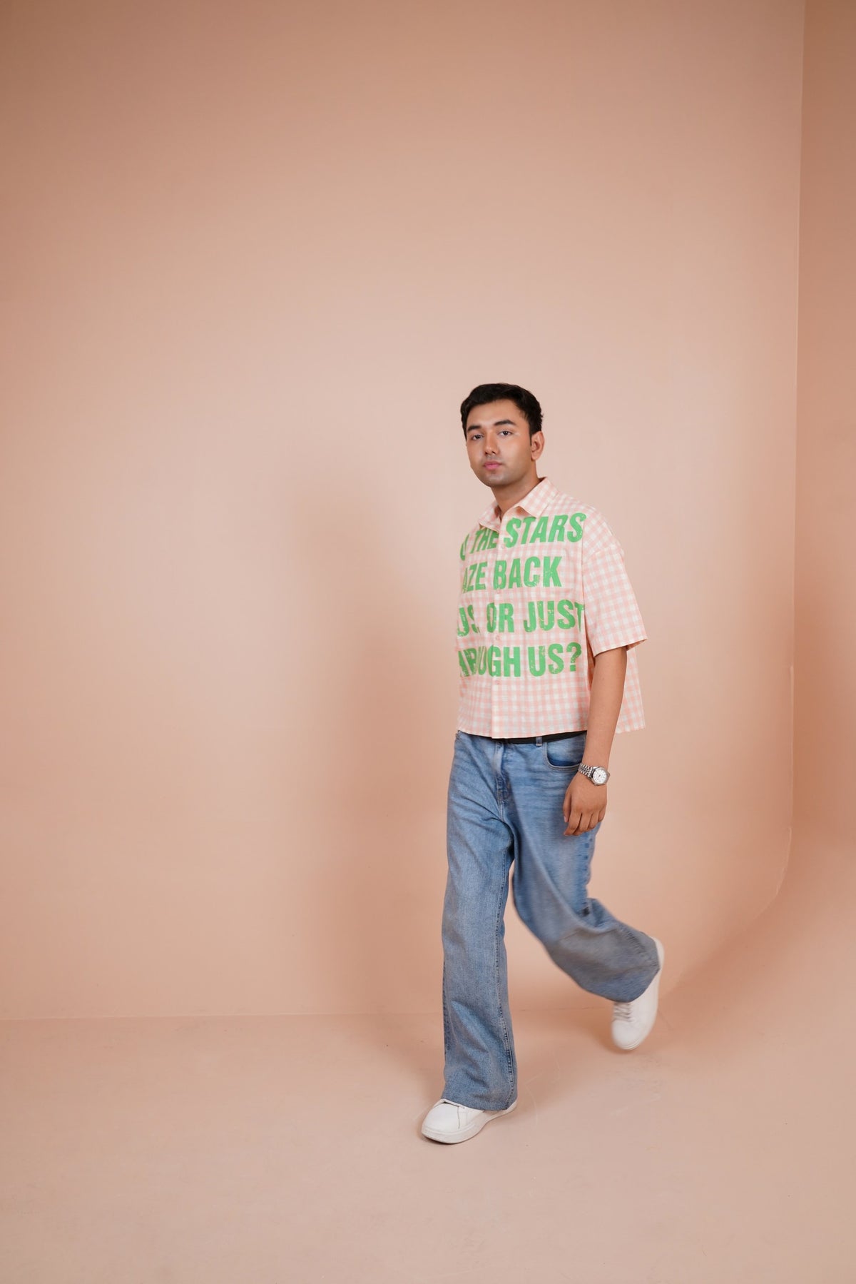 A man wearing a pink and white gingham short-sleeve t-shirt with green text that reads 'THE STARS ARE BACK OR JUST SLEEPING US?' paired with light blue jeans and white sneakers, standing against a solid peach-pink studio backdrop.