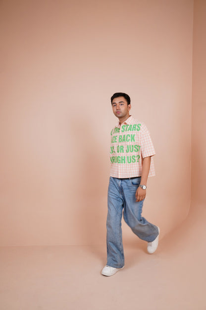 A man wearing a pink and white gingham short-sleeve t-shirt with green text that reads 'THE STARS ARE BACK OR JUST SLEEPING US?' paired with light blue jeans and white sneakers, standing against a solid peach-pink studio backdrop.