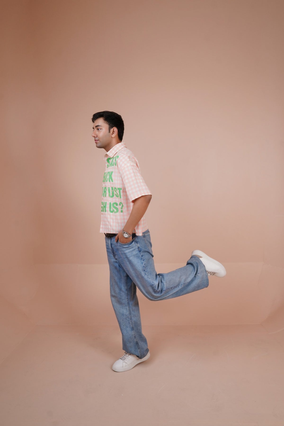 A man in a light pink and white checkered short-sleeve shirt with green text, blue jeans, and white sneakers, captured in a side profile pose against a solid peach-colored studio backdrop.
