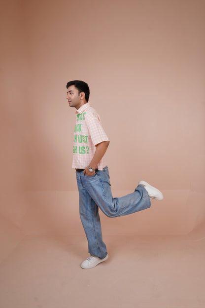 A man in a light pink and white checkered short-sleeve shirt with green text, blue jeans, and white sneakers, captured in a side profile pose against a solid peach-colored studio backdrop.